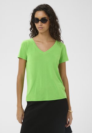 Femme aux cheveux bouclés portant des lunettes de soleil noires, un t-shirt vert vif à col en V, une jupe noire et un bracelet, debout devant un fond uni.