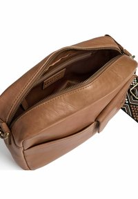 Brun läder crossbody-väska med dragkedjestängning, med en texturerad rem och en intern etikett. Detalj med ytterficka är synlig.