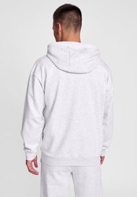 Haut de survêtement gris clair avec une grande capuche, des poignets côtelés et une taille côtelée. Texture douce avec une coupe surdimensionnée, mettant en avant un design simple et décontracté.