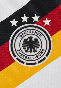 Schwarzer Adler-Emblem des Deutschen Fußball-Bundes mit vier Sternen darüber, auf einem strukturierten Stoffhintergrund in Schwarz-, Rot- und Gelbstreifen.