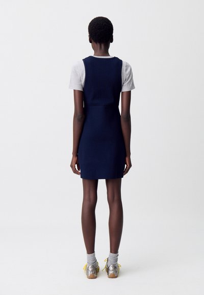 Robe marine avec un corsage côtelé, associée à un T-shirt gris. Le modèle porte des chaussures argentées avec des accents jaunes et des chaussettes grises.