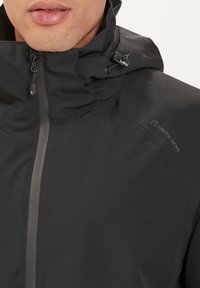 Sort vandtæt jakke med højt krave, lynlås foran og justerbare snore. Har et diskret logo på brystet og struktureret stof.