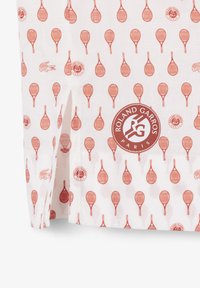 Weißer Stoff mit einem Muster aus roten Tennisschlägern und Roland-Garros-Logos, versehen mit einem großen, runden Roland-Garros-Paris-Emblem nahe dem unteren Rand.