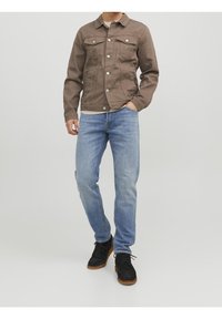 Giacca di cotone marrone con bottoni in metallo, due tasche sul petto, abbinata a jeans azzurri chiari e scarpe nere con suole marroni.