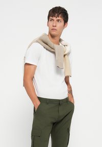 Weißes T-Shirt unter einem hellbeige- und cremefarbenen Schal. Olivgrüne Cargo-Hosen mit seitlichen Taschen. Modell steht mit den Händen in den Taschen.