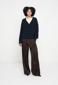 Cardigan bleumarin tricotat, cu decolteu în V, nasturi în față și buzunare frontale, asortat cu pantaloni maro largi și pantofi negri cu vârf ascuțit.