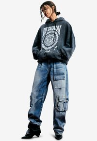 Czarna bluza z kapturem z białym nadrukowanym logo i grafiką, zestawiona z niebieskimi luźnymi dżinsami z wieloma kieszeniami i suwakami, noszona z czarnymi szpilkami.