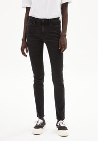 Jean skinny en denim noir avec une taille mi-haute, cinq poches et un effet légèrement délavé. Associé à des baskets noires et blanches, avec des lacets blancs.
