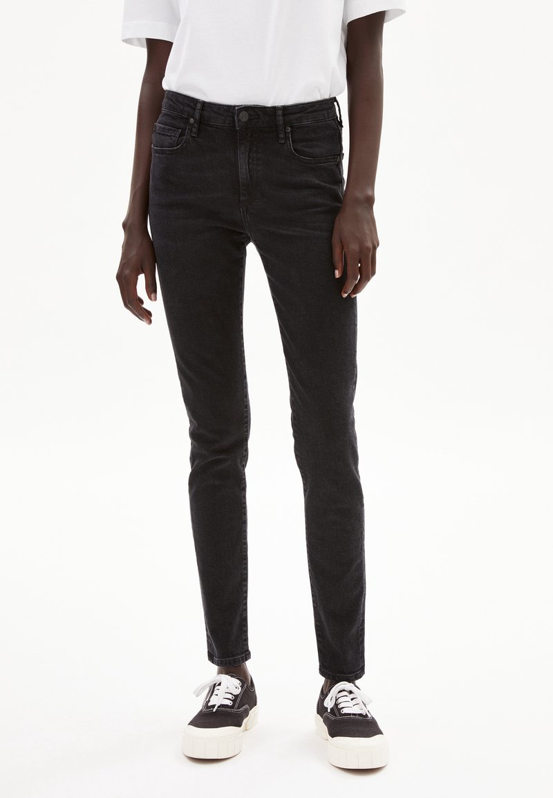 Jean skinny en denim noir avec une taille mi-haute, cinq poches et un effet légèrement délavé. Associé à des baskets noires et blanches, avec des lacets blancs.