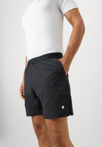 Homem vestido com calções de desporto pretos e camisa polo branca, de pé com uma mão no bolso, contra um fundo simples.