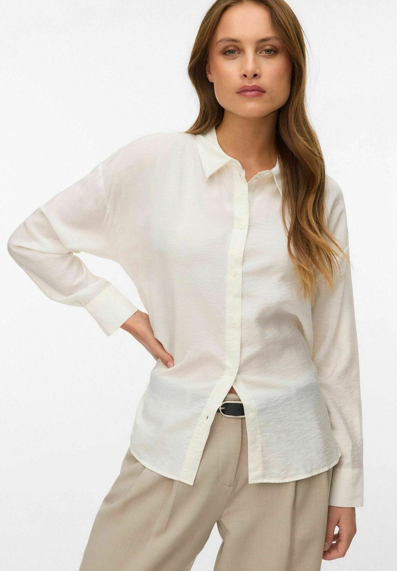 Femme aux cheveux longs portant une blouse crème boutonnée et un pantalon beige, debout avec une main sur la hanche, sur un fond uni.