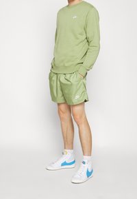 Olivově zelená mikina Nike s kulatým výstřihem, spárovaná s odpovídajícími lehkými šortkami. Bílé logo Nike a tenisky s vysokou obcí s modrými akcenty.