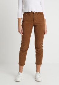 Pantalon en velours côtelé marron avec une coupe slim, longueur cropped et poches avant, associé à un haut blanc à manches longues et des chaussures blanches.