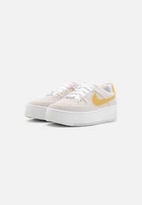 Białe zamszowe sneakersy z grubą białą podeszwą. Żółte akcenty na pięcie i logo Nike. Sznurowane białymi sznurowadłami, z perforowanymi detalami.