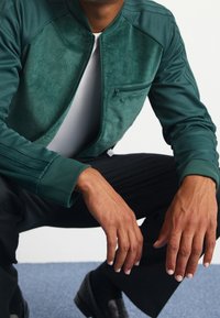 Teal kordjacka i manchester med ribbade ärmar och ficka med dragkedja. Matchas med svarta byxor och svarta skor. Bakgrunden har en blå matta.