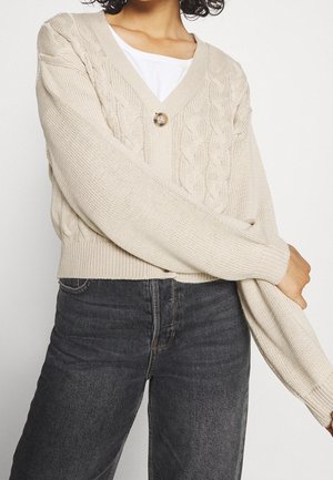Femme portant un cardigan beige à torsades avec un seul bouton sur une chemise blanche et un jean taille haute gris foncé sur un fond uni.