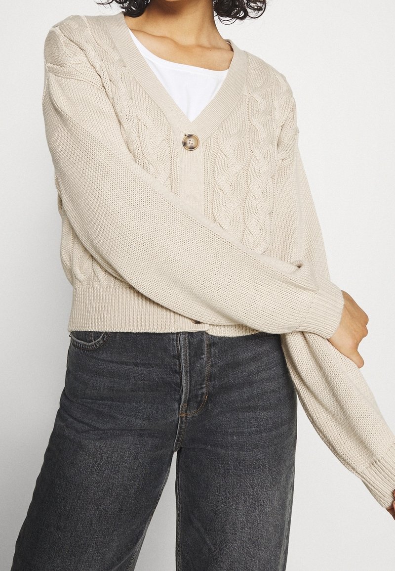 Femme portant un cardigan beige à torsades avec un seul bouton sur une chemise blanche et un jean taille haute gris foncé sur un fond uni.