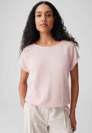Femme aux longs cheveux bouclés portant un haut en maille rose clair à manches courtes et un pantalon crème taille haute, debout devant un fond uni.