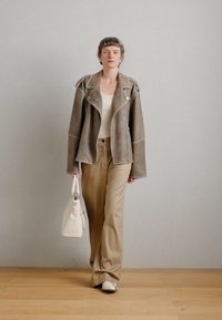 Brūna zamšādas jaka ar baltu šuvumu, balta topiņa, bēšu plašu bikses un krēmkrāsas somiņa. Grīda ir gaiši koka. Fons ir neitrāls.