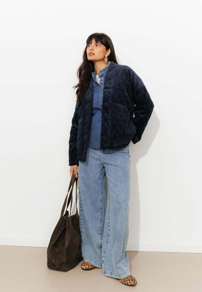 Veste matelassée bleu marine avec une surface texturée, associée à un haut en denim, un jean à jambe large et des chaussures à imprimé léopard. Sac fourre-tout marron foncé.