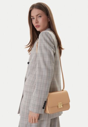 Donna con capelli castani lisci che indossa un completo grigio a quadri e porta una borsa a tracolla in pelle beige con fibbia dorata.