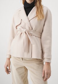 Nainen, joka pukeutunut beigeen villatakkiin, jossa on vyö, mustan pooloneuleen ja vaaleiden beigenväristen cordurohousujen päälle, seisoo yksinkertaisen taustan edessä.