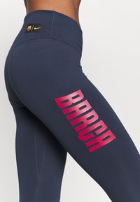 Marina leggings med hög midja, tillverkade av stretchigt material, med stora rosa "BARCA" bokstäver på vänstra låret och en Nike-logga på midjebandet.