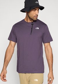 Μωβ κοντομάνικη μπλούζα από βαμβάκι, με μικρό λευκό λογότυπο στο στήθος. Φορεμένη με μπεζ παντελόνια και μαύρο καπέλο τύπου bucket.