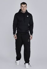 Melna kapuce ar priekšējo kabatu un logo uz krūšu, kombinēta ar melniem cargo biksēm ar sānu kabatām un logo detaļu. Baltas kedas.