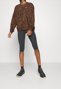 Bruin luipaardprint sweatshirt met lange mouwen, gecombineerd met zwarte knielange legging en zwarte sneakers met groene accenten.
