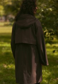 Cappotto impermeabile marrone scuro con cappuccio, caratterizzato da un design cropped, maniche lunghe e una texture liscia. Nessun fissaggio o motivo visibile.
