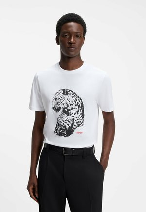 Mand iført en hvid t-shirt med et sort-hvidt leopardmotiv og teksten "HUGO", kombineret med sorte bukser og bælte, stående.