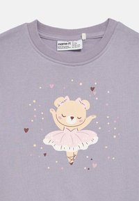 Ljubičasta majica dugih rukava s ilustracijom slatkog medvjedića u ružičastoj tutu suknji. Dizajn je ukrašen zvjezdicama i srcima u svijetlo ružičastoj i smeđoj boji.