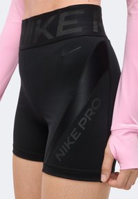 Gros plan d'une personne portant un short Nike Pro noir taille haute avec le logo et la marque Nike, associé à un haut à manches longues rose clair.