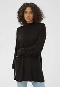 Pulover negru cu mâneci lungi și guler înalt, realizat dintr-un material moale și neted. Are o croială lejeră și crăpături laterale la tiv. Purta peste blugi deschis la culoare.