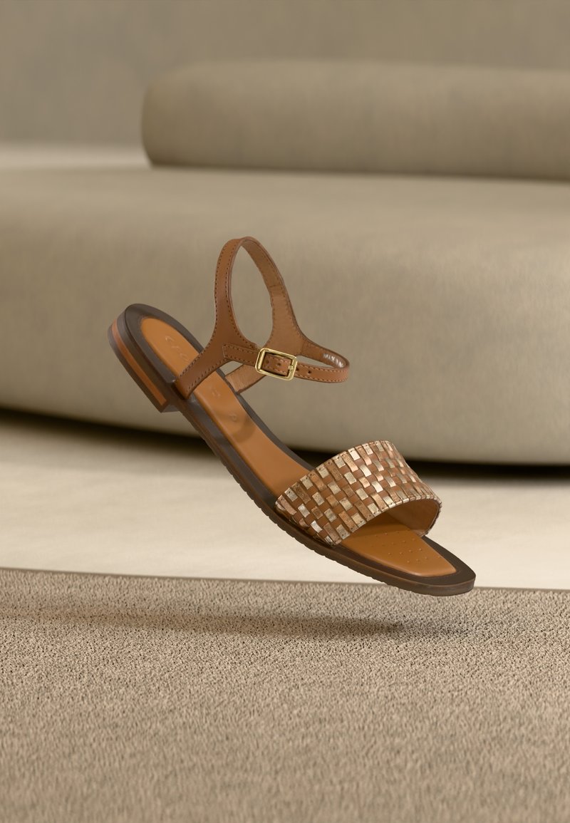Hnědá kožená sandále s tkaným kovovým páskem, nastavitelnou přezkou na kotníku, plochou podrážkou a texturovanou stélkou. Vznáší se nad neutrálním povrchem.