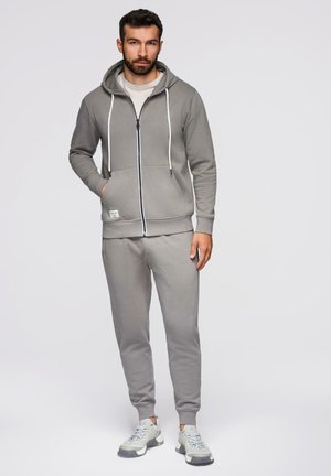 Mand iført grå lynlås-hættetrøje og matchende træningsbukser med hvide sneakers, stående foran en ensartet lys baggrund.