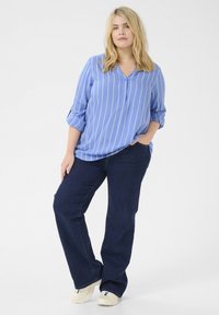 Blaue Hemd mit weißen vertikalen Streifen, kombiniert mit dunklen Jeans mit weitem Bein und hellen Sneakers. Lässiger Schnitt und entspannter Stil.