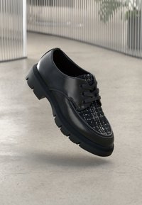 Chaussure noire à lacets avec un cou-de-pied en cuir brillant et un bout en tissu texturé, dotée d'une semelle épaisse et robuste et de accents contrastés.