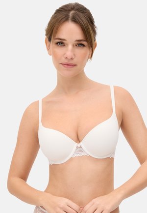 Mujer joven con un sujetador blanco liso con aro y ribete de encaje, de pie frente a un fondo claro y simple.