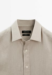 Leinenhemd in einem hellbeigen Farbton mit klassischem Kragen, fünf Knöpfen und einer strukturierten Stoffoberfläche. Markenlabel innen am Kragen sichtbar.