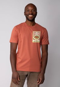 Een roestkleurige katoenen t-shirt met een grafisch ontwerp dat pastabelichting en de tekst "DE PASTA" bevat. Korte mouwen en een ronde hals.