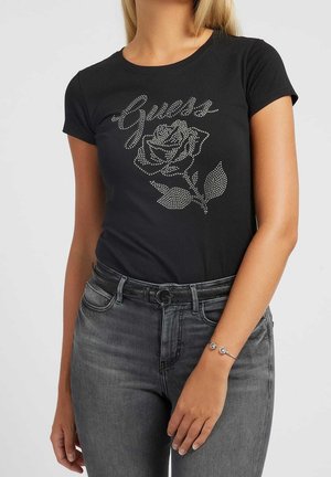 Black cotton t-shirt featuring a rhinestone rose design and "Guess" text. Pairing gray jeans with a belt is also visible.