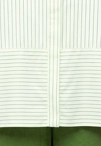 Tissu blanc avec de fines rayures verticales et horizontales foncées et une couture verticale centrale sur un tissu vert en dessous.