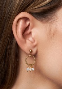 Guldörhängen med en texturerad cirkulär design och tre hängande vita pärlor. Örhängena bärs i ett piercingöra.
