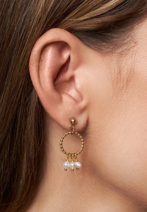 Los pendientes de aro dorados presentan un diseño circular texturizado con tres perlas blancas colgantes. Los pendientes se llevan en una oreja perforada.