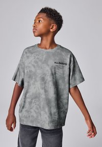 Chlapec s krátkymi kučeravými vlasmi, oblečený v sivom tie-dye tričku AllSaints a tmavých džínsach, pozerá sa bokom s mierne rozpaženými rukami.