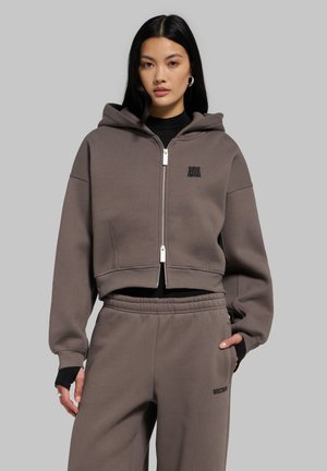 Jeune femme aux longs cheveux noirs porte un sweat à capuche marron zippé et un pantalon de survêtement assorti sur un fond gris uni.