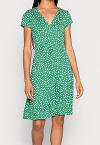 Zielona sukienka z krótkim rękawem i dekoltem w kształcie litery V, zdobiona białym wzorem w kropki, z dopasowaną talią i rozkloszowanym dołem. Wykonana z lekkiego materiału.