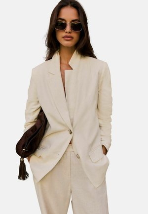 Frau in beigem Anzug und Sonnenbrille, hält eine braune Handtasche mit Quasten unter dem Arm und steht mit den Händen in den Taschen.